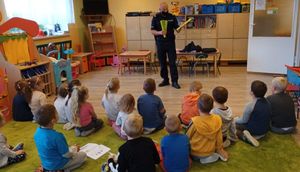 Umundurowany policjant stoi przed grupą przedszkolaków. Dzieci siedzą na podłodze. Policjant prezentuje trzymane w rękach żółte opaski odblaskowe.