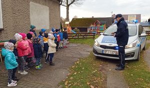 Dzieci stoją przy elewacji przedszkola. Przed nimi stoi policyjny radiowóz z włączoną sygnalizacją świetlną, a przy nim umundurowany policjant, który opowiada o służbie.