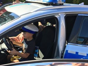 Dwoje dzieci siedzi w radiowozie, na pierwszym planie na fotelu kierowcy siedzi chłopczyk w czapce policyjnej.