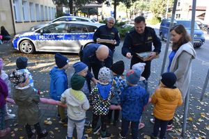 Policjant i strażnik miejski wręczają dzieciom odblaski. W tle policyjny radiowóz. Dzieci stoją w szeregu.
