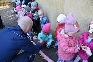 Policjant przyczepia odblask na rękawie kurtki małej dziewczynki w różowej czapce. Obok inne dzieci stoją w szeregu.