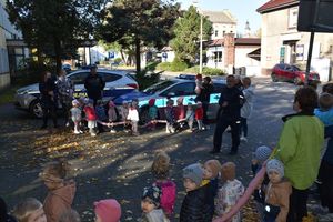 Policjant prowadzi pogadankę. Dzieci stoją po dwóch stronach w szeregu. W tle radiowóz policji i straży miejskiej.