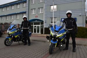 Dwaj policjanci w białych hełmach ochronnych stoją obok nowych motocykli. W tle wejście do oleskiej komendy Policji.