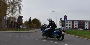 Policjant jadący na motocyklu na łuku drogi. W tle budynek mieszkalny.