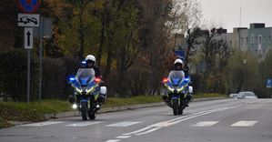 Dwaj policjanci na motocyklach jadą ulica Klonową w Oleśnie. Motocykle maja załączone światła błyskowe uprzywilejowania. W tle bloki mieszkalne.