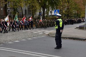 Przemarsz uczestników uroczystości ulicą Pieloka w Oleśnie. Na skrzyżowaniu z ulicą Krasickiego policjant ruchu drogowego zabezpiecza przemarsz.