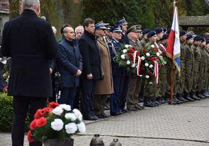 Uczestnicy uroczystości stoją w szeregu przy pomniku Lotników Polskich. Na pierwszym planie wiązanka biało-czerwonych kwiatów leżąca na płycie pomnika.