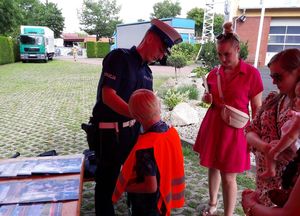 Policjant ruchu drogowego pomaga ubrać chłopcu pomarańczową kamizelkę odblaskową. Obok stoją dwie kobiety. Jedna trzyma na rękach małe dziecko.