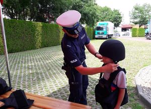 Policjant ruchu drogowego pomaga założyć małemu chłopcu czarny hełm ochronny. Chłopiec ma na sobie granatową kamizelkę kuloodporną z napisem POLICJA.