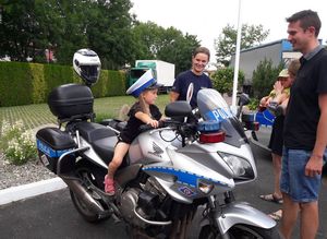 Mała dziewczynka w policyjnej czapce z białym pokrowcem siedzi na policyjnym motocyklu. Po lewej stronie stoi policjantka ruchu drogowego, a prze motocyklem rodzice dziewczynki.