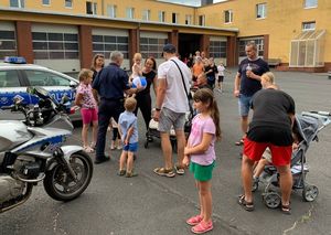 Grupa ludzi, dorosłych i dzieci. Po lewej stronie policyjny motocykl i radiowóz. Policjant podaje dzieciom niebiesko-białą piłkę plażową.
