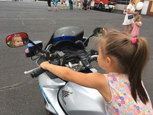 Mała dziewczynka w sukience na ramiączkach w kolorowe kwiatuszki siedzi na policyjnym motocyklu. W lewym lusterku odbicie jej twarzy. W tle inni ludzie i wóz bojowy straży pożarnej.