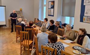 Sala kominkowa MDK w Oleśnie, seniorze siedzą przy stole, około 16 osób, za nimi na końcu stołu stoi policjant obok niego ekran z prezentowanym slajdem.