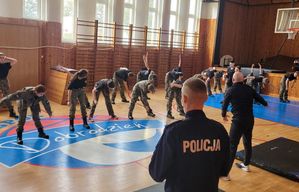 Sala gimnastyczna. Na pierwszym planie policjant w granatowym mundurze ćwiczebnym z napisem na plecach POLICJA. W tle instruktor i ćwicząca przed nim grupa uczniów.