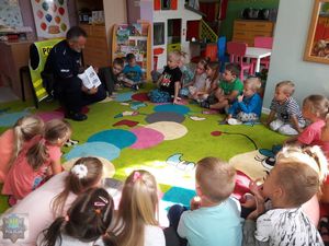 Policjant prezentuje dzieciom książeczkę &quot;Bezpieczny przedszkolak&quot; - dzieci siedzą na dywanie.