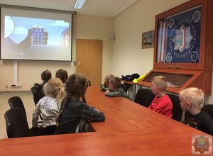 dzieci siedzą w sali konferencyjnej KPP w Oleśnie - oglądają prezentację na temat bezpieczeństwa na ekranie slajd z numerem alarmowym 112. Po prawej stronie gablota ze sztandarem oleskiej komendy.