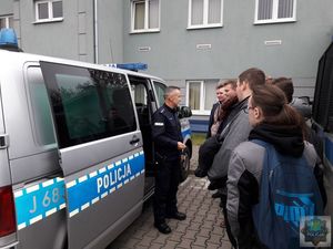 Plac zewnętrzny KPP w Oleśnie uczniowie stoją z boku policyjnego VW T6 , umundurowany funkcjonariusz stoi przy  otwartych drzwiach pojazdu, rozmawia z uczniami.